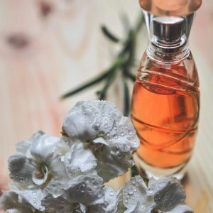 Glass perfume bottle alongside dew-kissed white carnation flowers on wood surface.