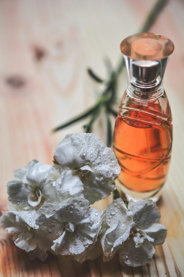 Glass perfume bottle alongside dew-kissed white carnation flowers on wood surface.